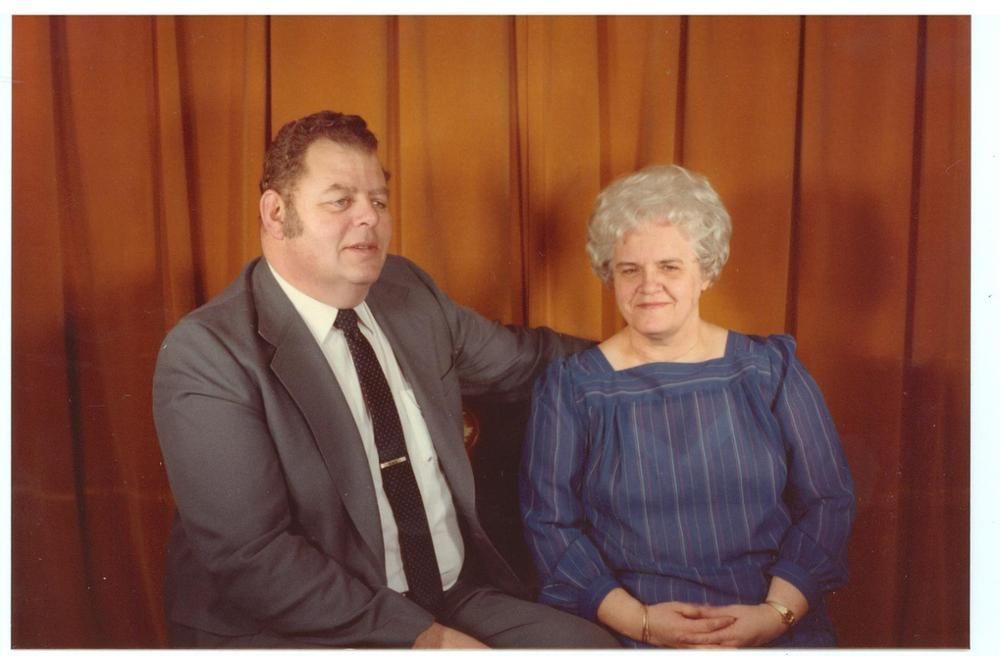 Two seated people in formal clothes against a wood-paneled wall, one resting a hand on the other's shoulder.