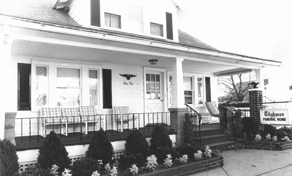 Black-and-white view of a porch-front house with a sign in the yard and flower beds along the walkway