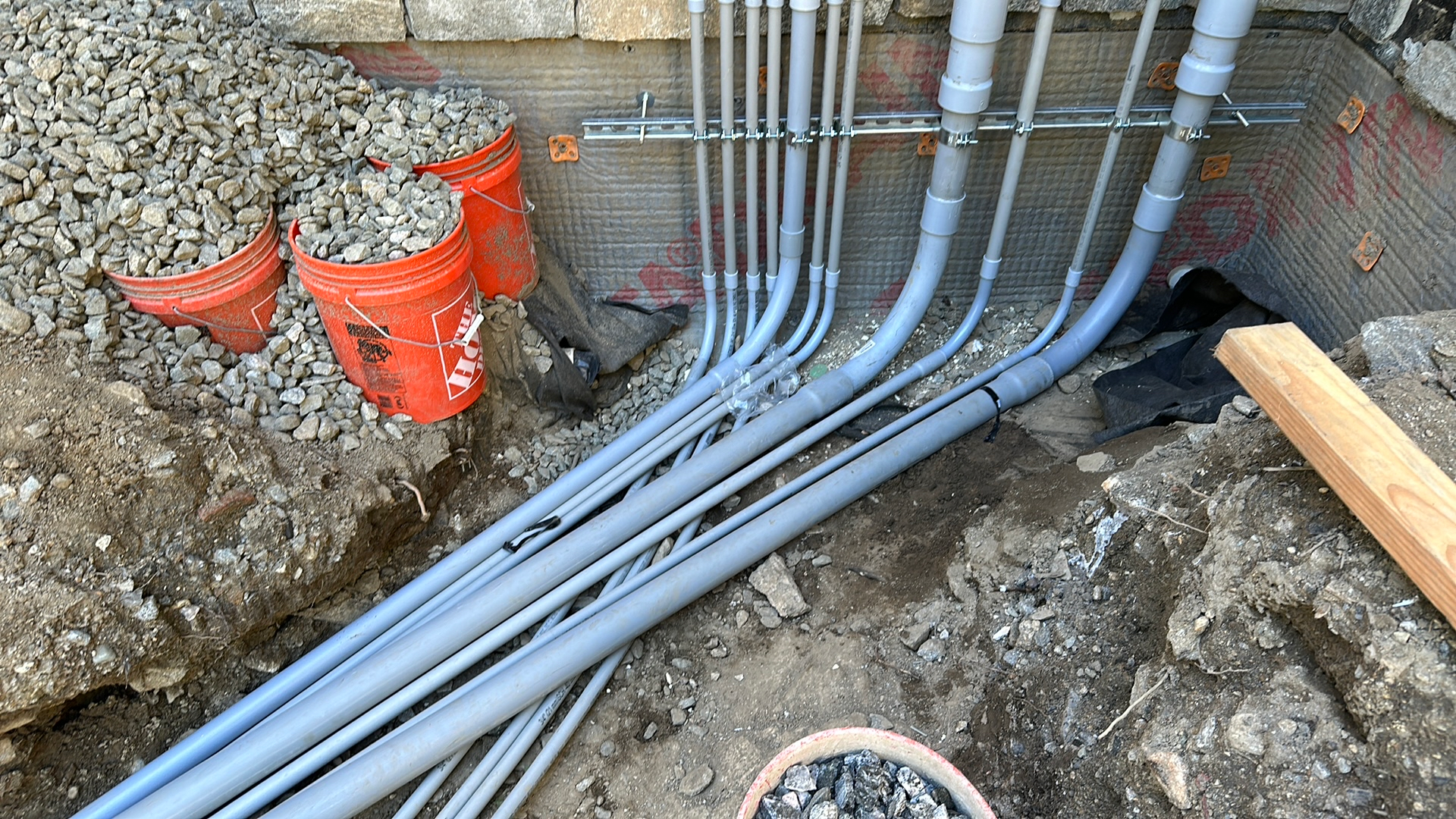Multiple grey PVC electrical conduits transition from wall-mounted struts into a trench in a dirt and gravel construction site.