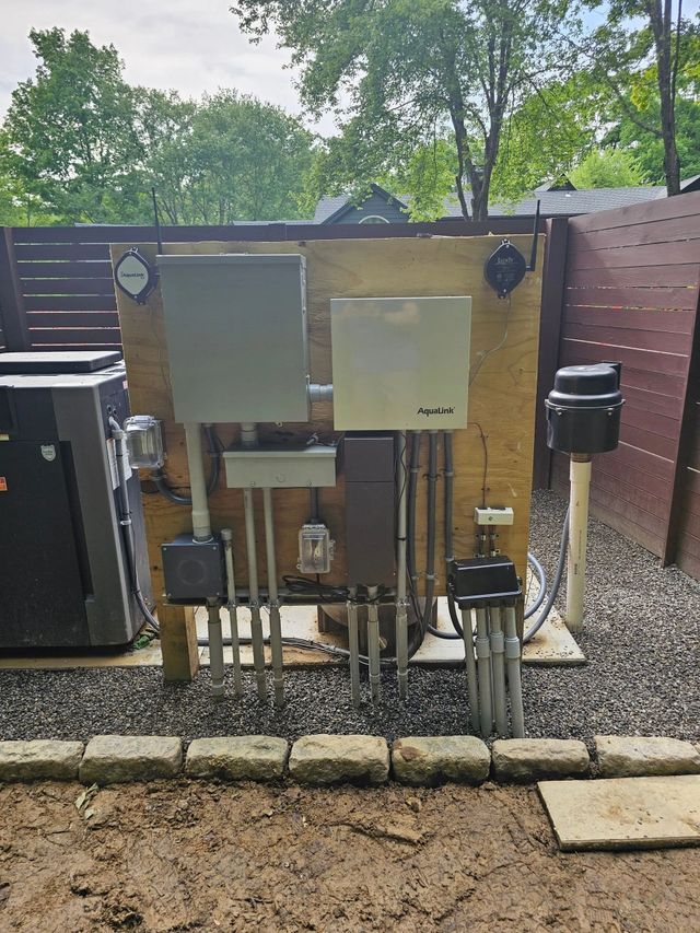 Electrical equipment, including a gray transfer switch and panel, mounted on a plywood board outdoors with conduit.