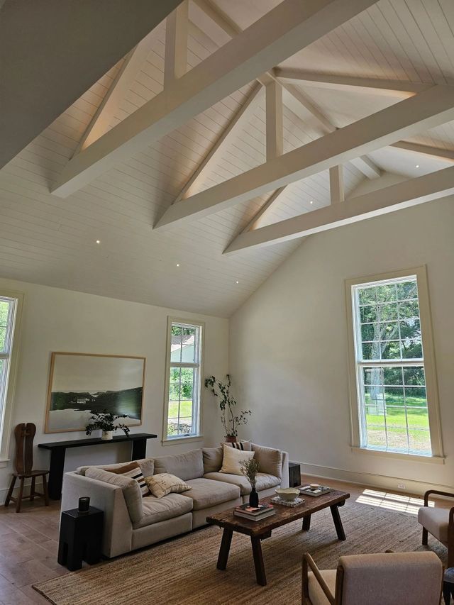 A bright, airy living room with a vaulted ceiling, exposed wooden trusses, cream walls, a large sectional, and windows.