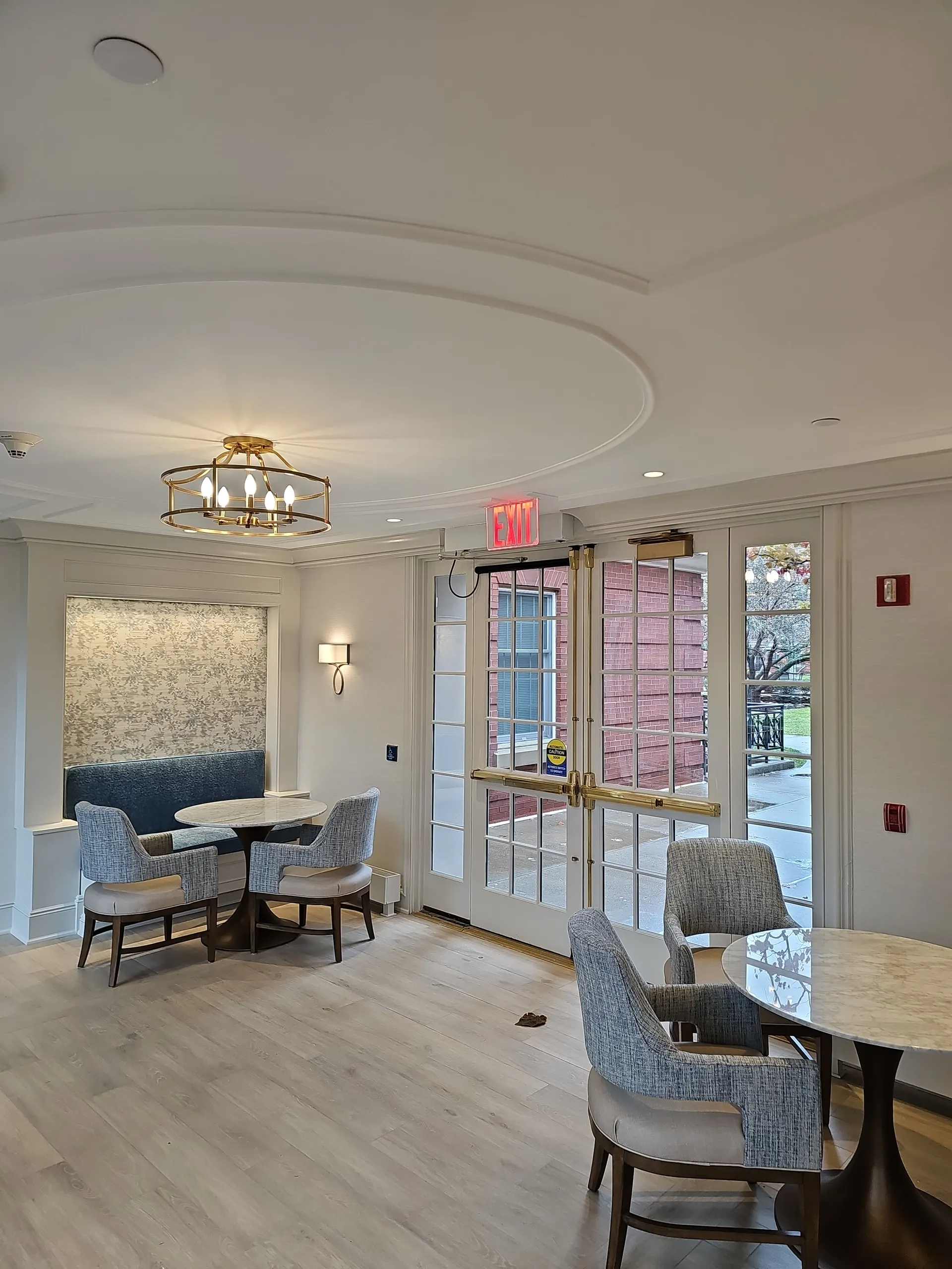 A well-lit room features two tables with patterned chairs, a decorative wall, and glass doors leading to the outdoors.