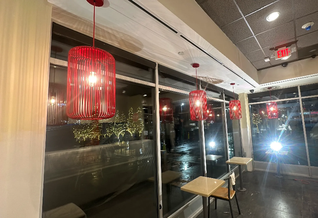 A row of cylindrical red pendant lights hangs in a modern, window-lined cafe with small tables and seating.