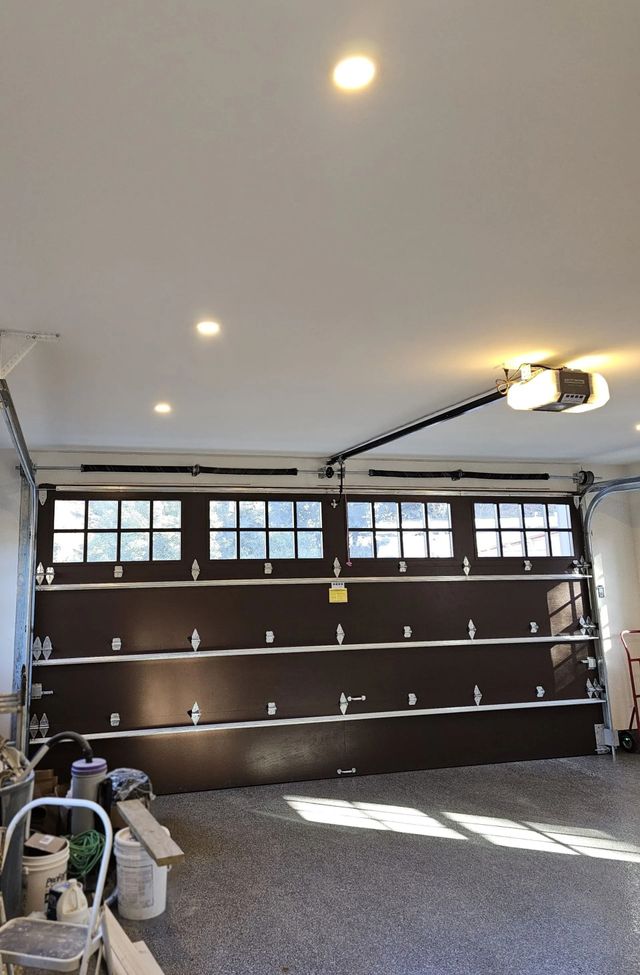 A dark brown, four-section garage door with small windows in the top panel, inside a garage with ceiling lights.