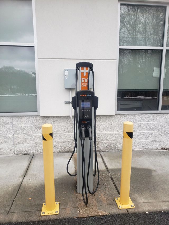 An electric vehicle charging station stands between two yellow bollards against a wall with windows.