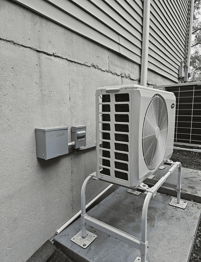 An outdoor HVAC heat pump unit sits on a metal stand against a concrete wall foundation under a house with vinyl siding.