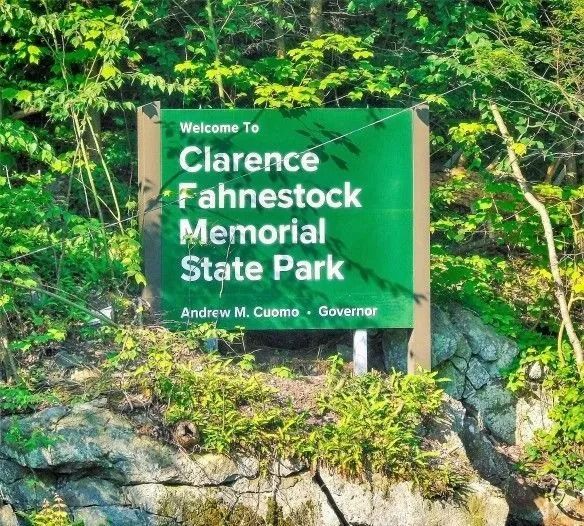 Green sign reading "Welcome To Clarence Fahnestock Memorial State Park" with a rocky, forest background.