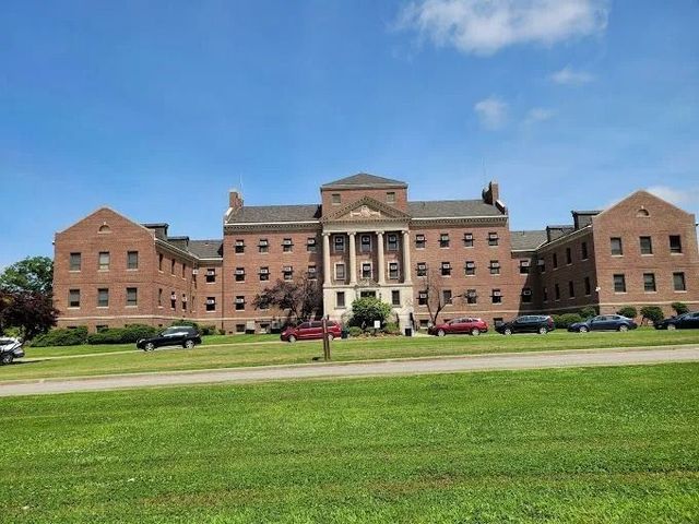 A multi-story brick building with a central columned entrance, surrounded by green lawns and parked cars under a blue sky.