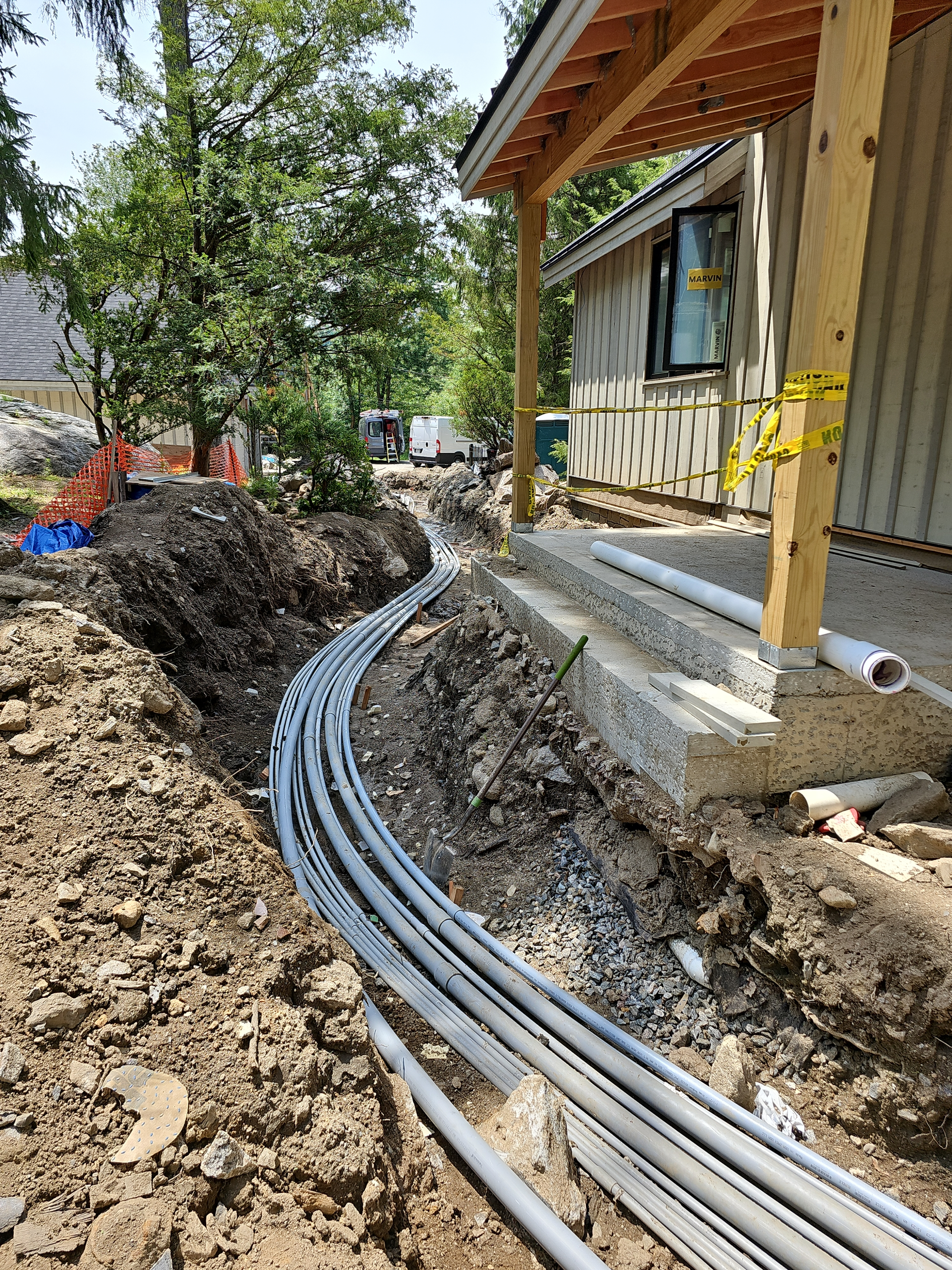 A trench beside a building frame containing multiple grey conduits leading towards the foundation.