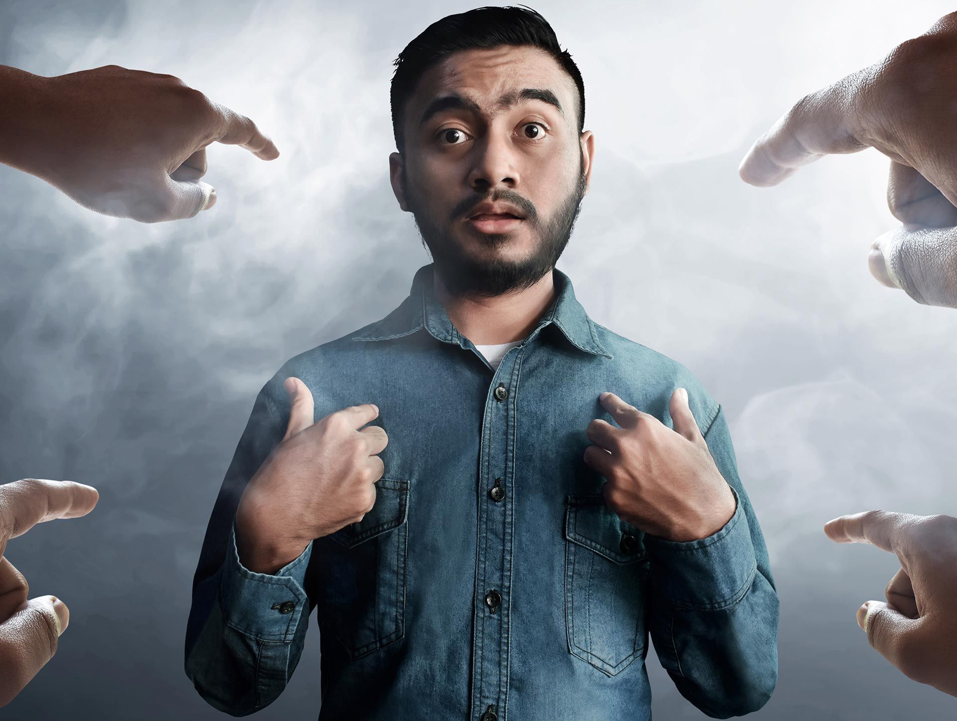 Confused man in a denim shirt pointing to himself, surrounded by large pointing hands in a smoky background Confused man in a denim shirt pointing to himself, surrounded by large pointing hands in a smoky background