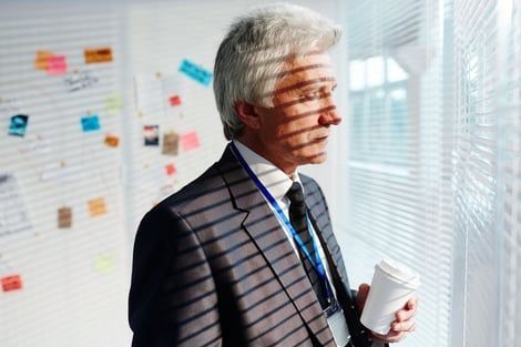 A person in a suit and lanyard holding a coffee cup gazes out a window with slatted blinds casting shadows on their face.