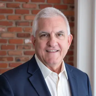A person with short grey hair, wearing a white shirt and navy blazer, smiles before a brown brick wall.