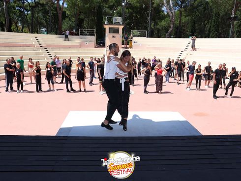 Due persone ballano su un palco all'aperto di fronte a una folla in una piazza soleggiata