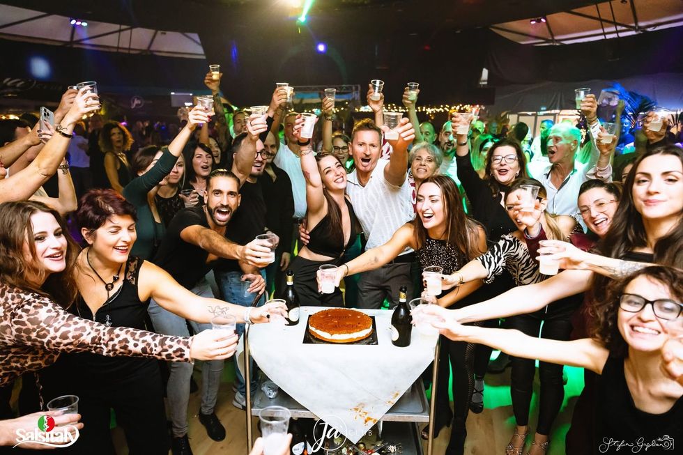Scena di festa affollata con persone che brindano intorno a una torta su un tavolo