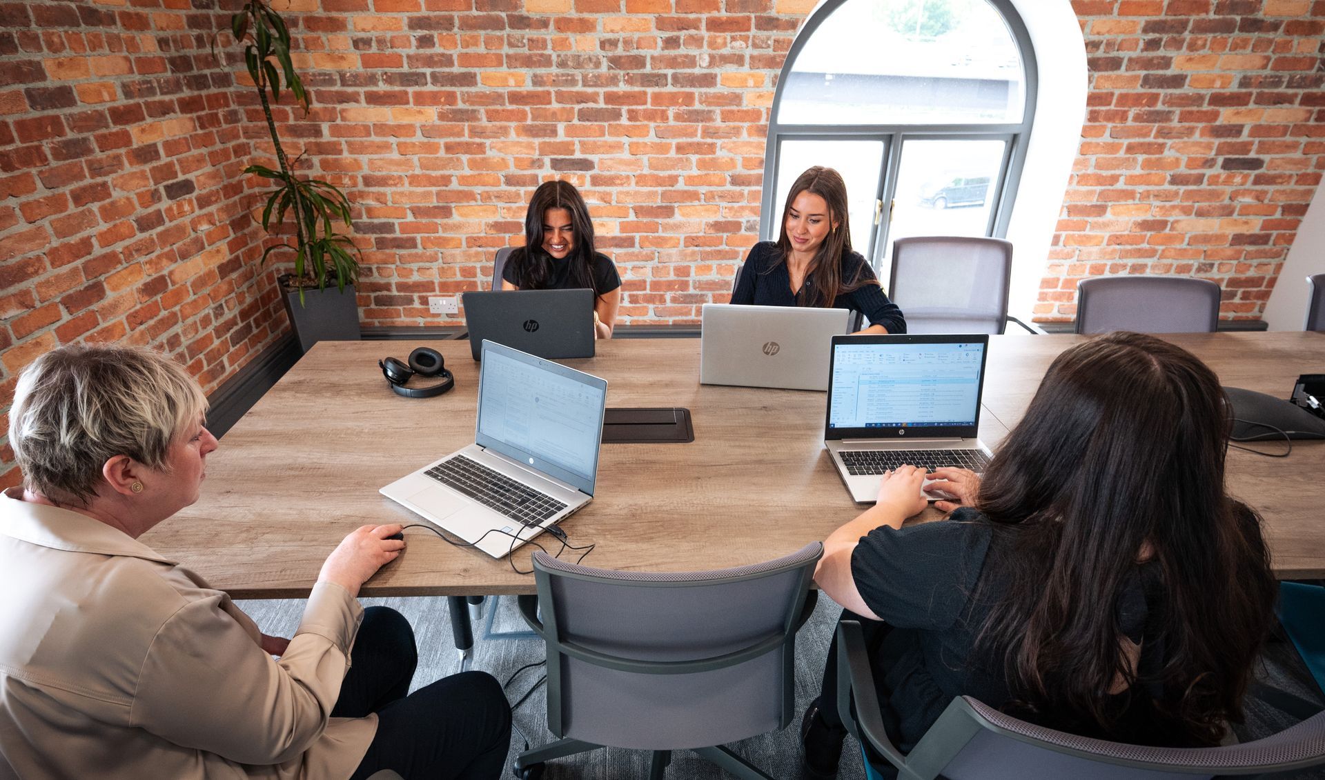 4 women sat at a large desk with laptops in front of them in an office with brick walls