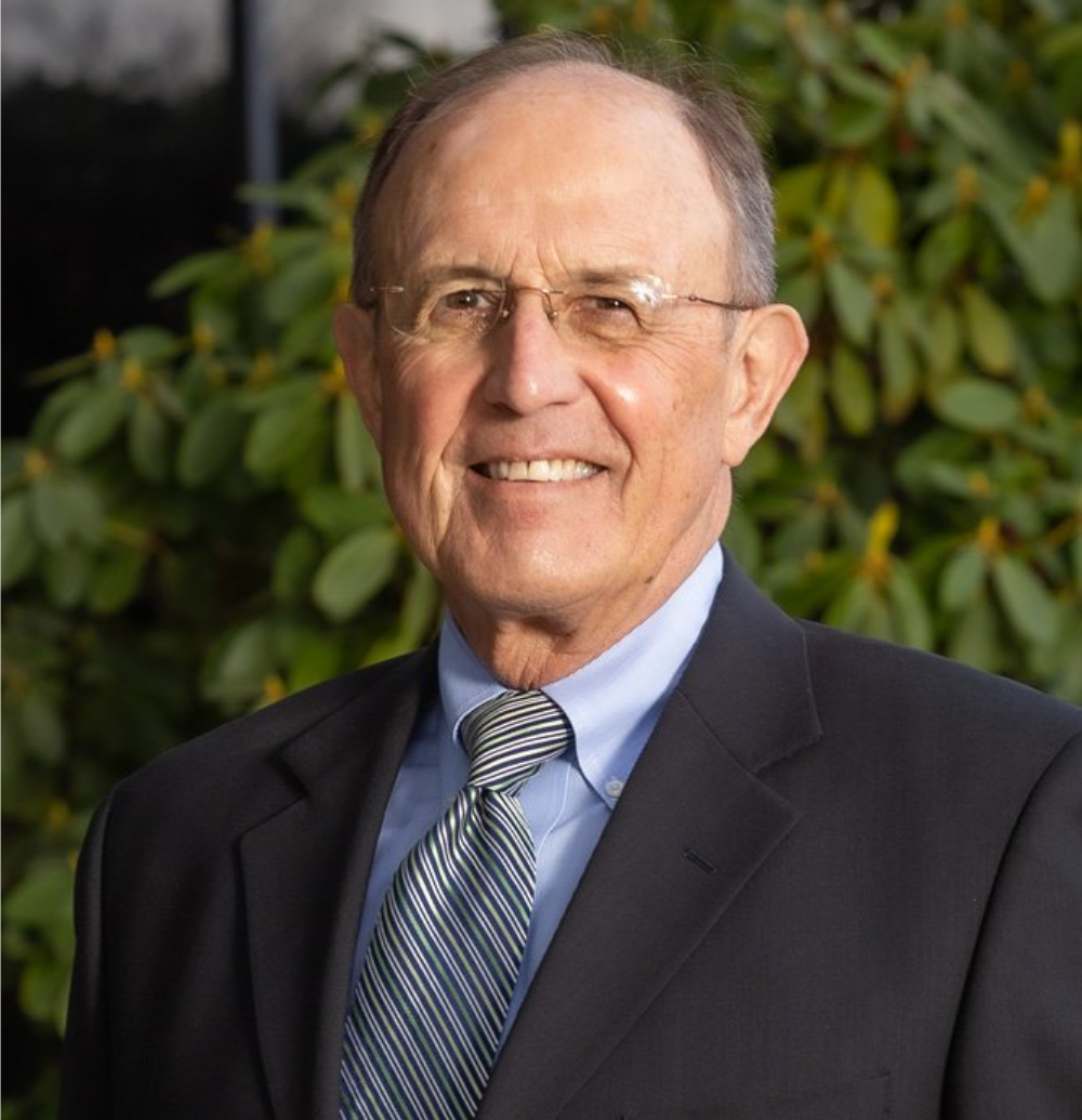Man wearing glasses and suit smiles in front of green foliage.