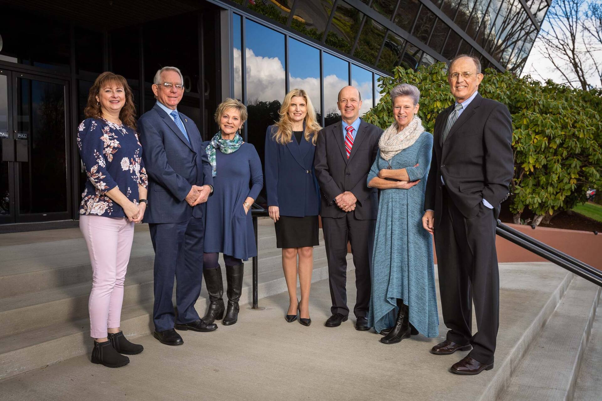 Group of seven people standing outside a building.  Two women and five men in business attire smiling.