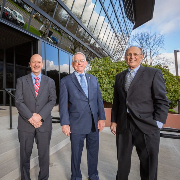 Three men in suits stand in front of a modern building with reflective glass windows.