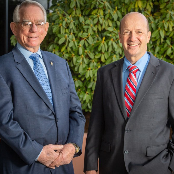 Two men in suits standing outside; one in blue, one in gray, both smiling.
