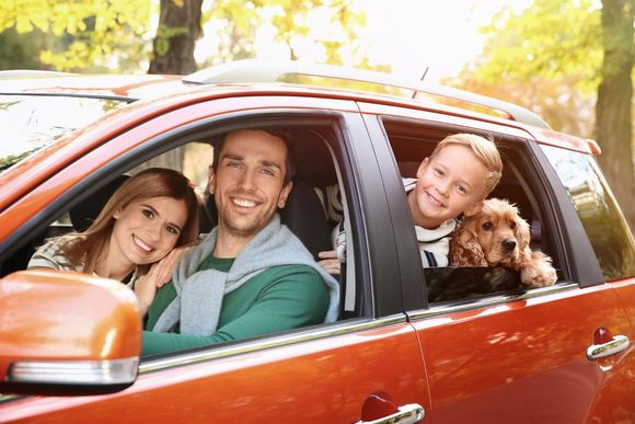 Family smiling in a red car, with a child and dog leaning out the back window.