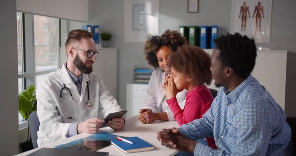 Doctor consulting with three patients at a clinic table, holding a tablet and discussing paperwork.