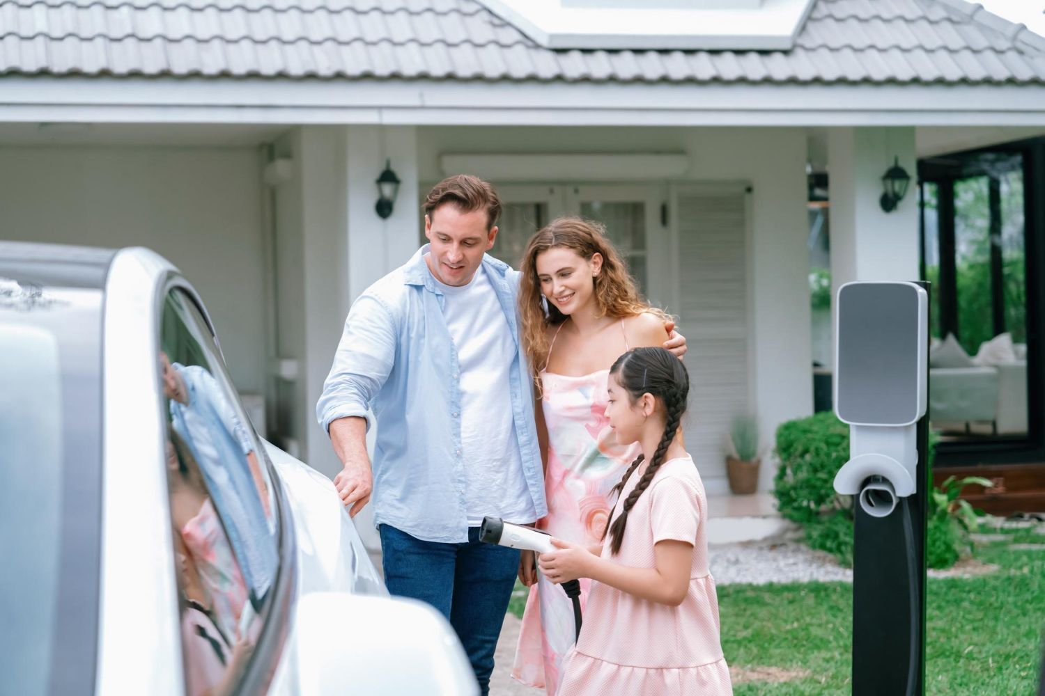 Family posing by a white car in a suburban driveway, with a home and charging station in the background.