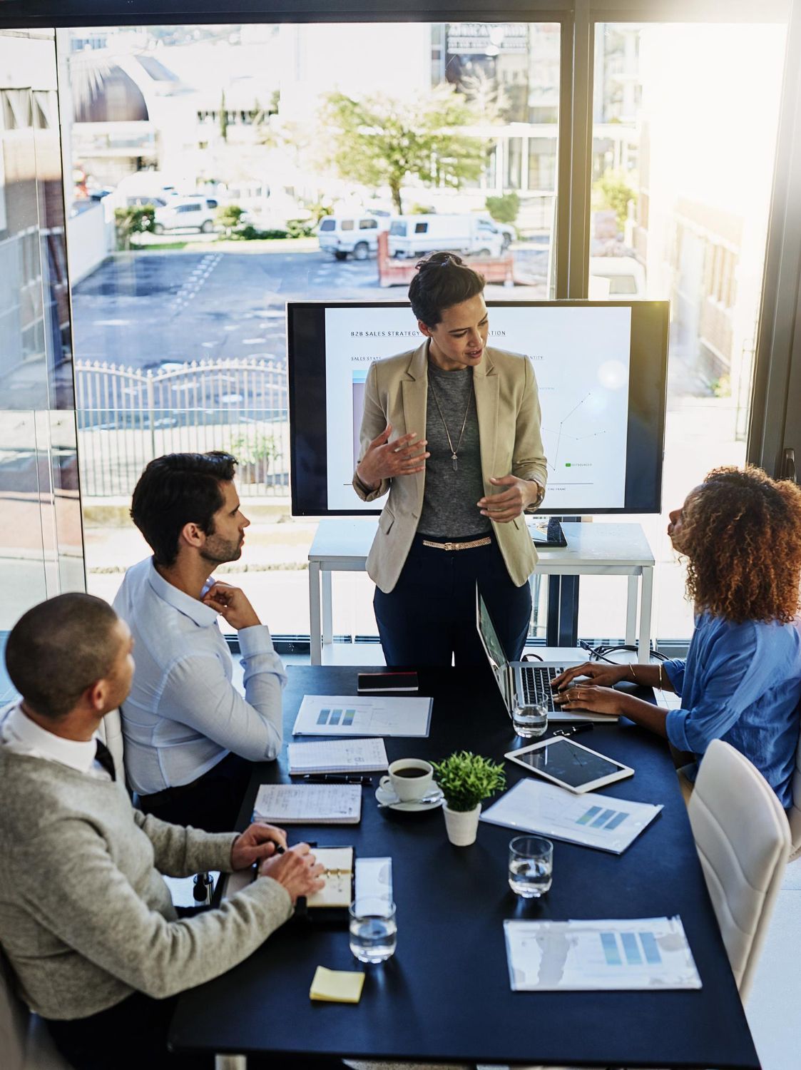 Business meeting in a bright office, with a presenter speaking beside a screen while colleagues take notes.