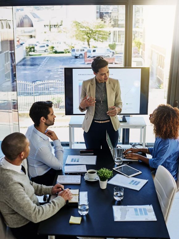Business meeting in a bright office, with a presenter speaking beside a screen while colleagues take notes.