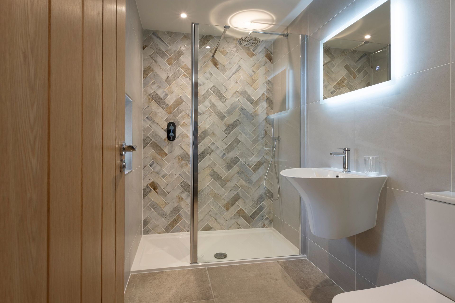 Modern bathroom with glass shower, herringbone tile, wall sink, and illuminated mirror.