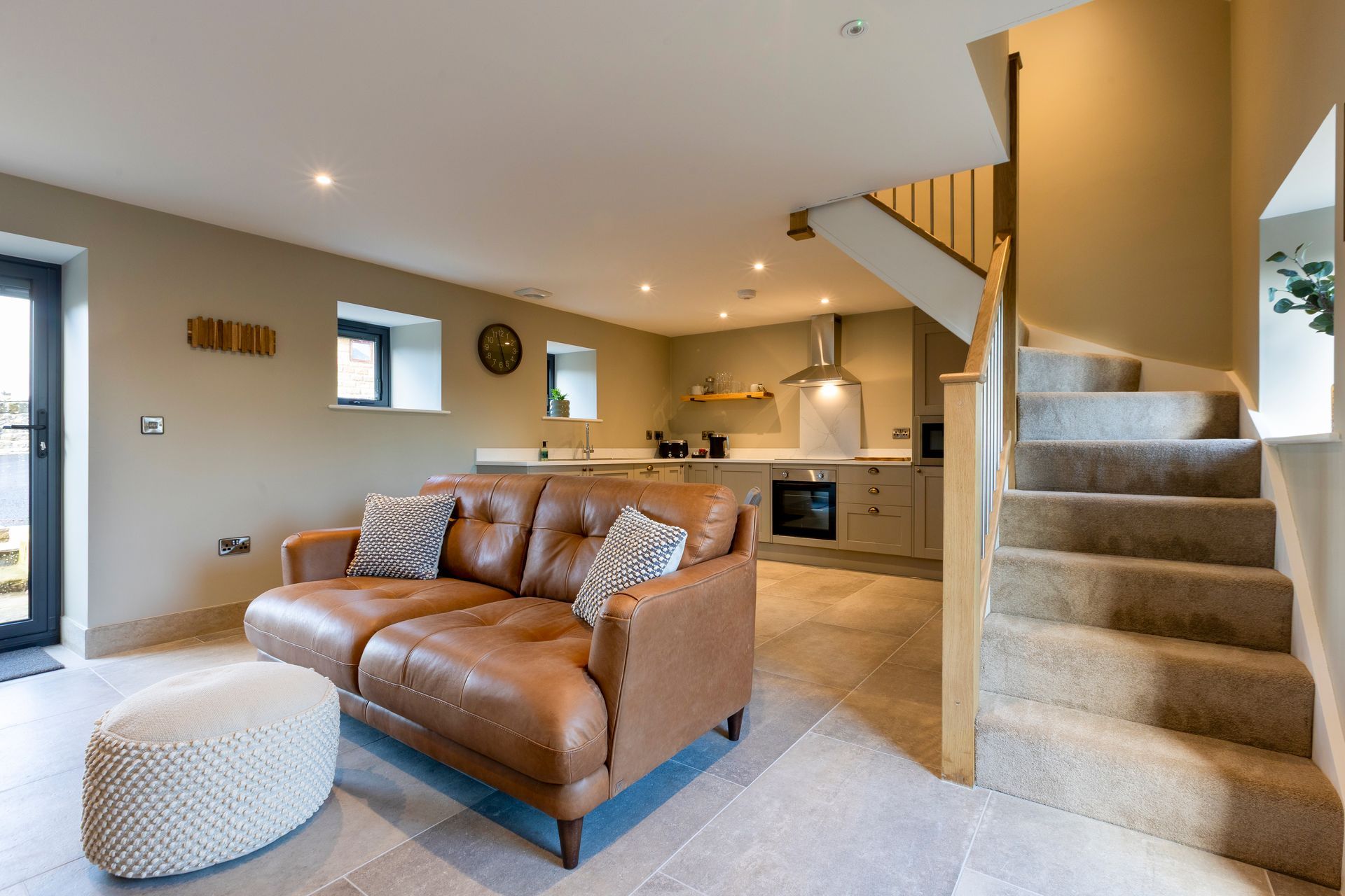 Modern living room with brown leather sofa, carpeted stairs, and open kitchen with warm lighting