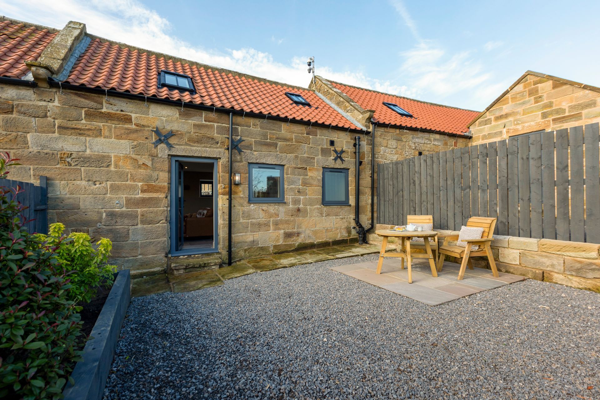 Stone courtyard with gravel, wooden table and chairs, and a rustic cottage with red-tiled roof.
