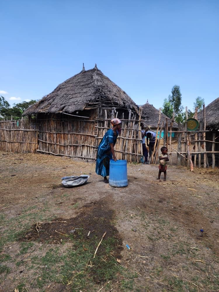 A woman is standing next to a blue barrel in front of a thatched hut.