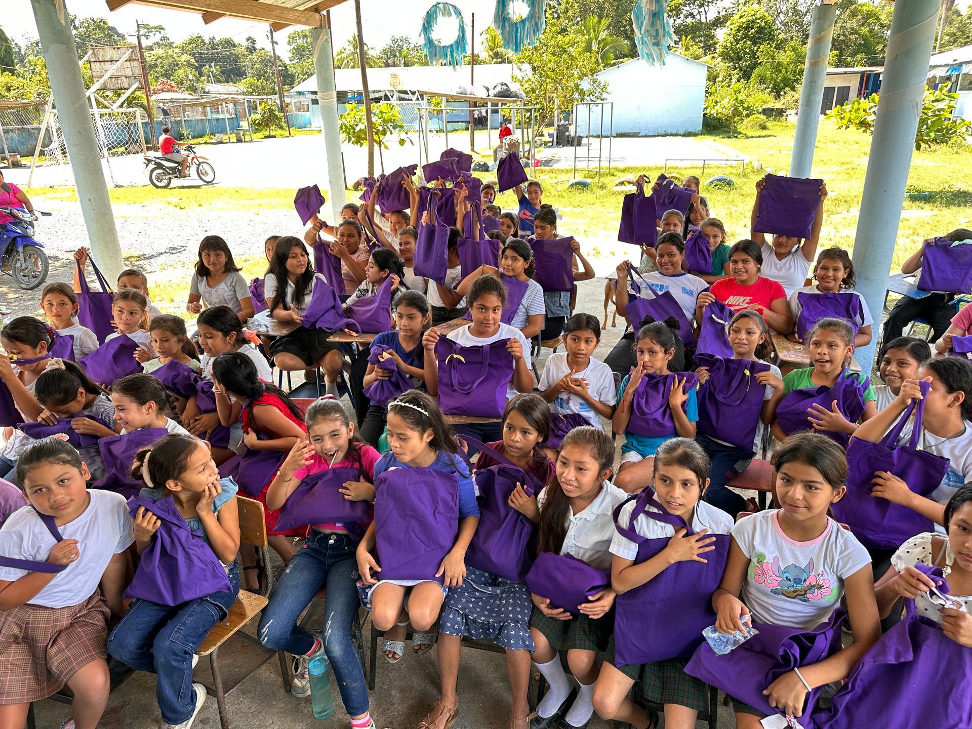 A large group of children are sitting under a canopy holding purple bags.