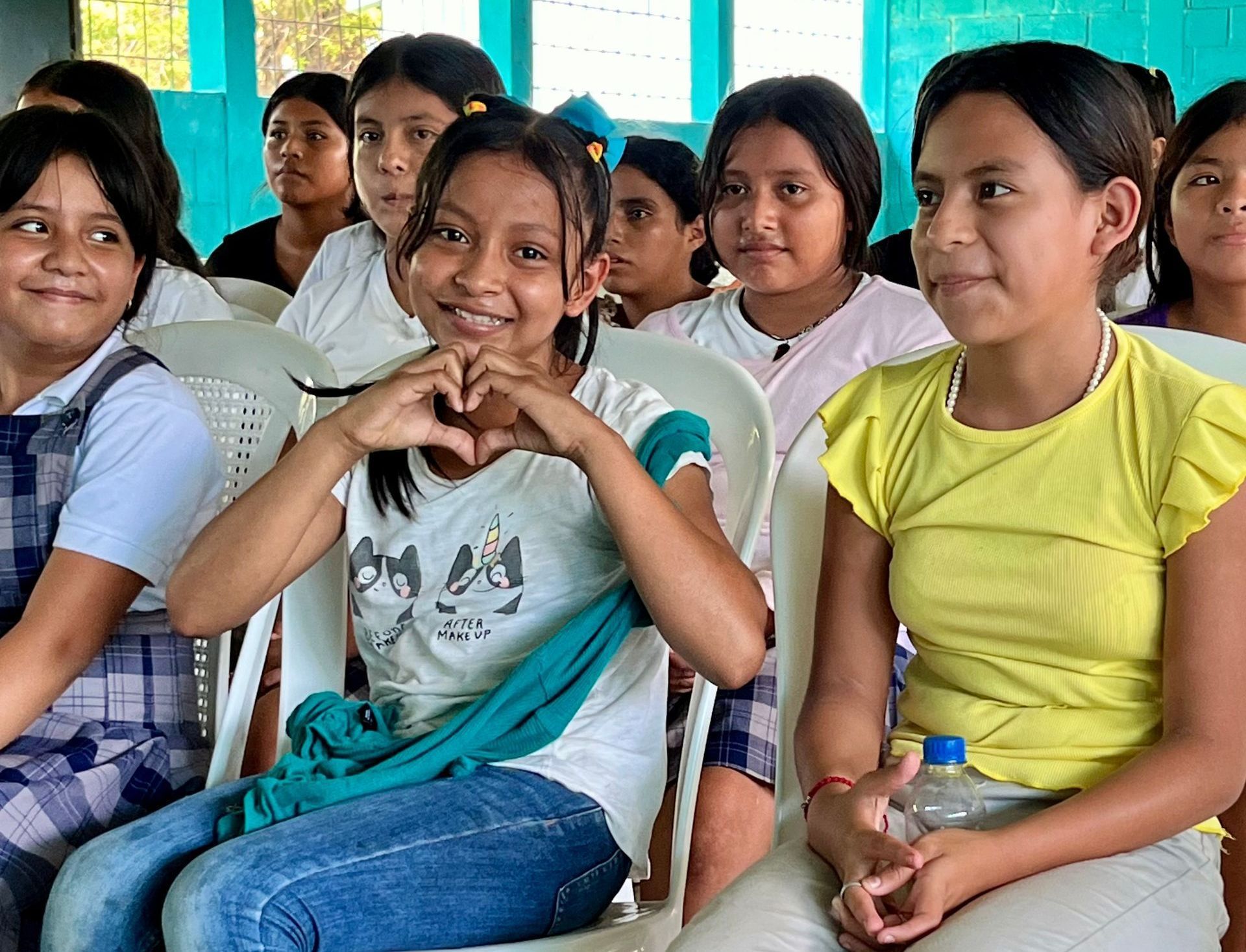 A group of young girls are sitting in chairs and one girl is making a heart shape with her hands.