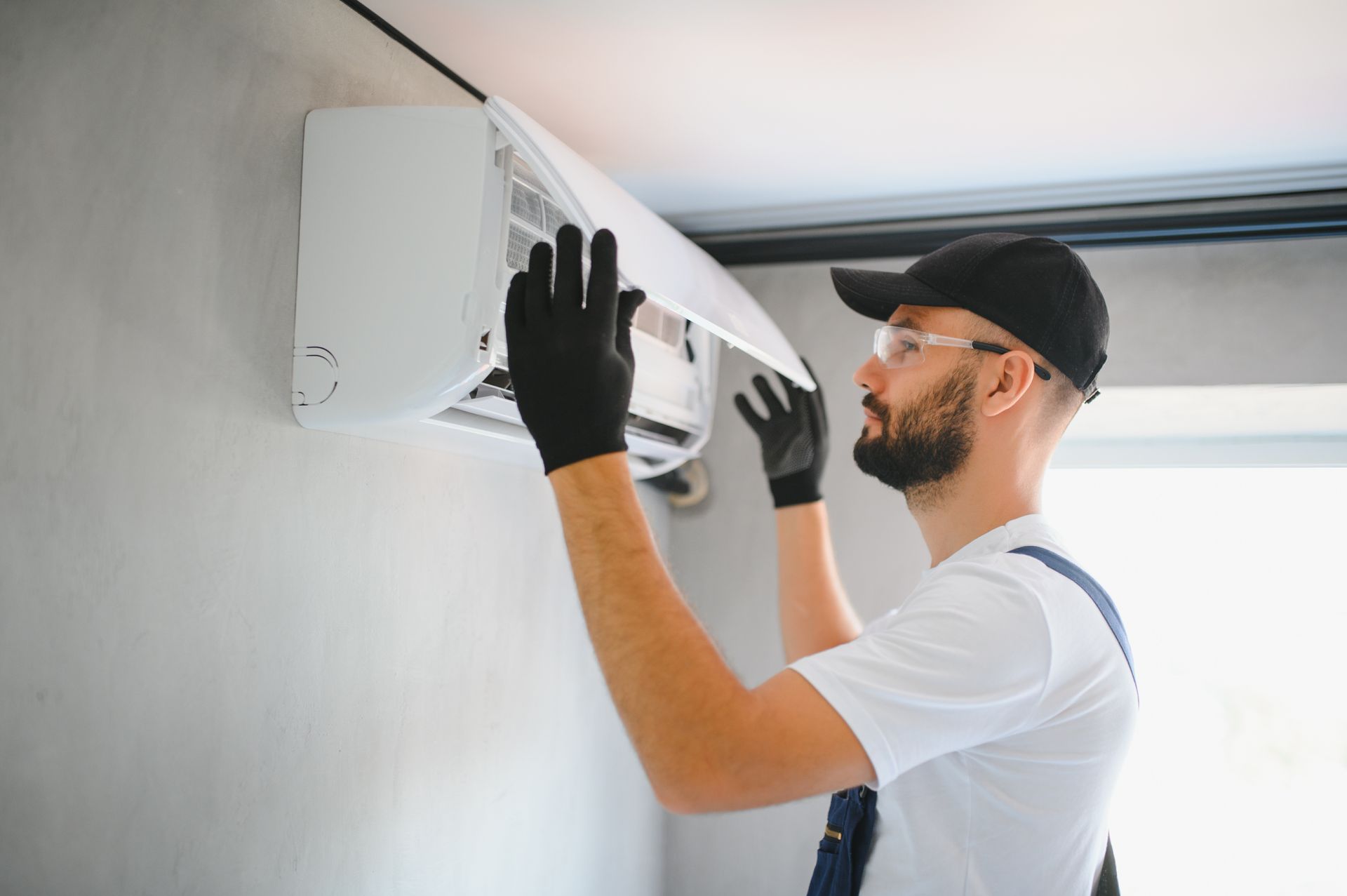A technician in protective eyewear and gloves performs maintenance on a wall-mounted air conditioner.