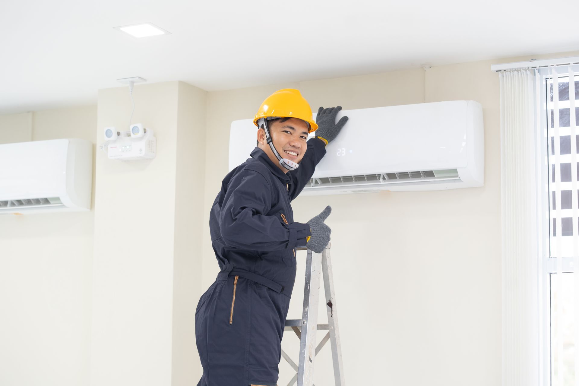 A technician in a hard hat and uniform on a ladder, smiling and giving a thumbs-up while working on a wall-mounted AC unit.