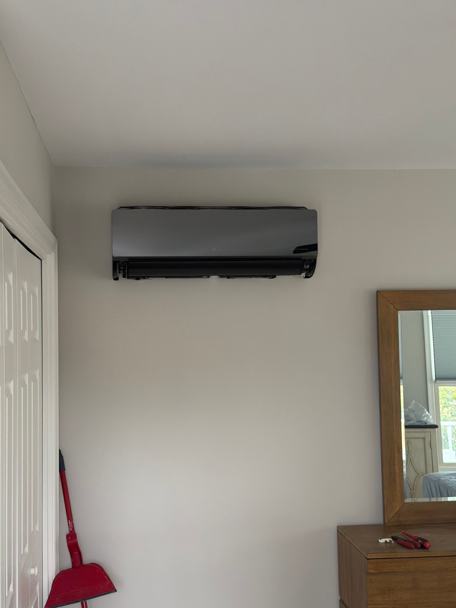 Wall-mounted air conditioner above a dresser in a plain bedroom with a broom and mirror nearby