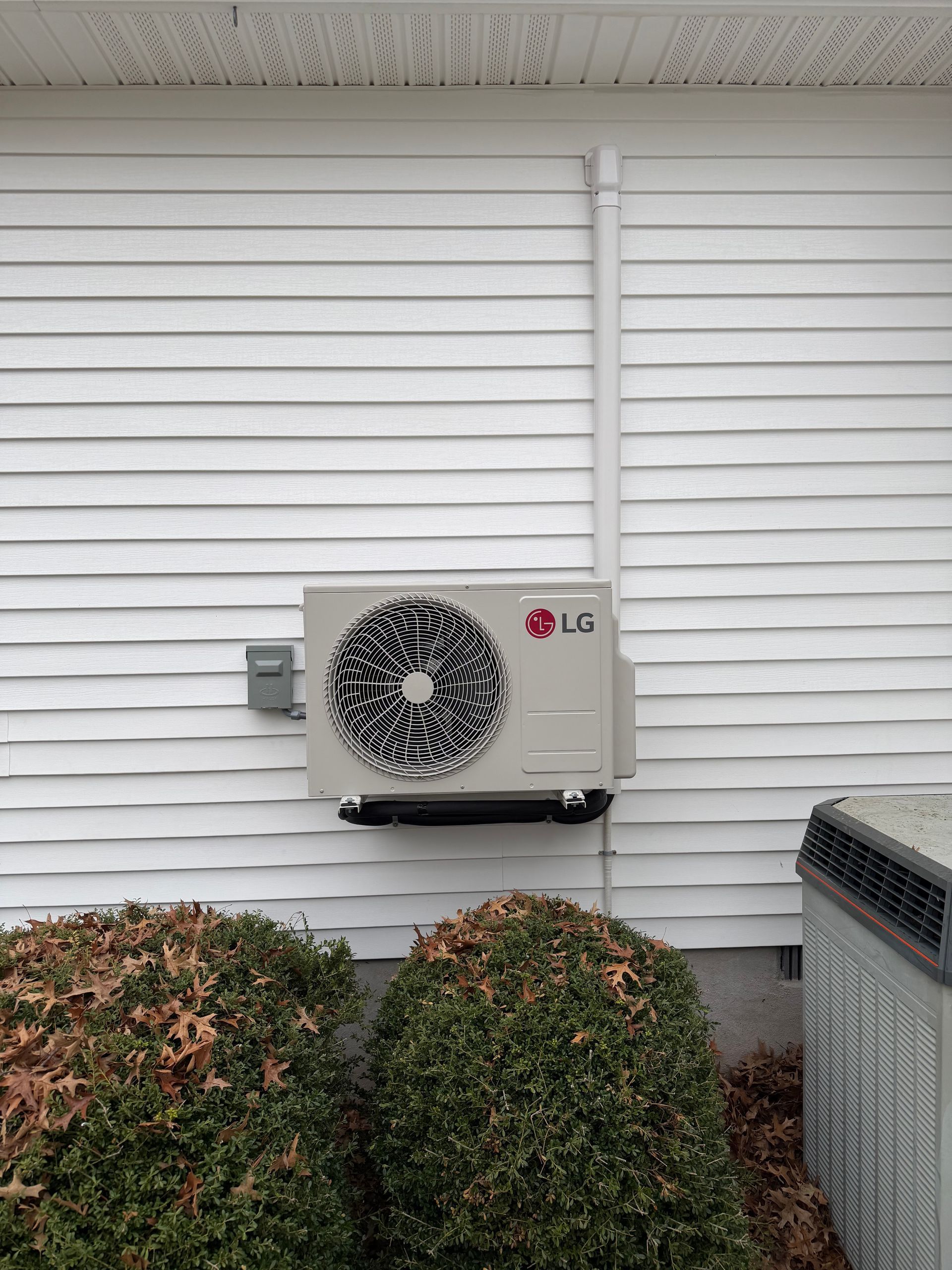 Outdoor HVAC unit mounted on white siding beside shrubs and a woven outdoor bin