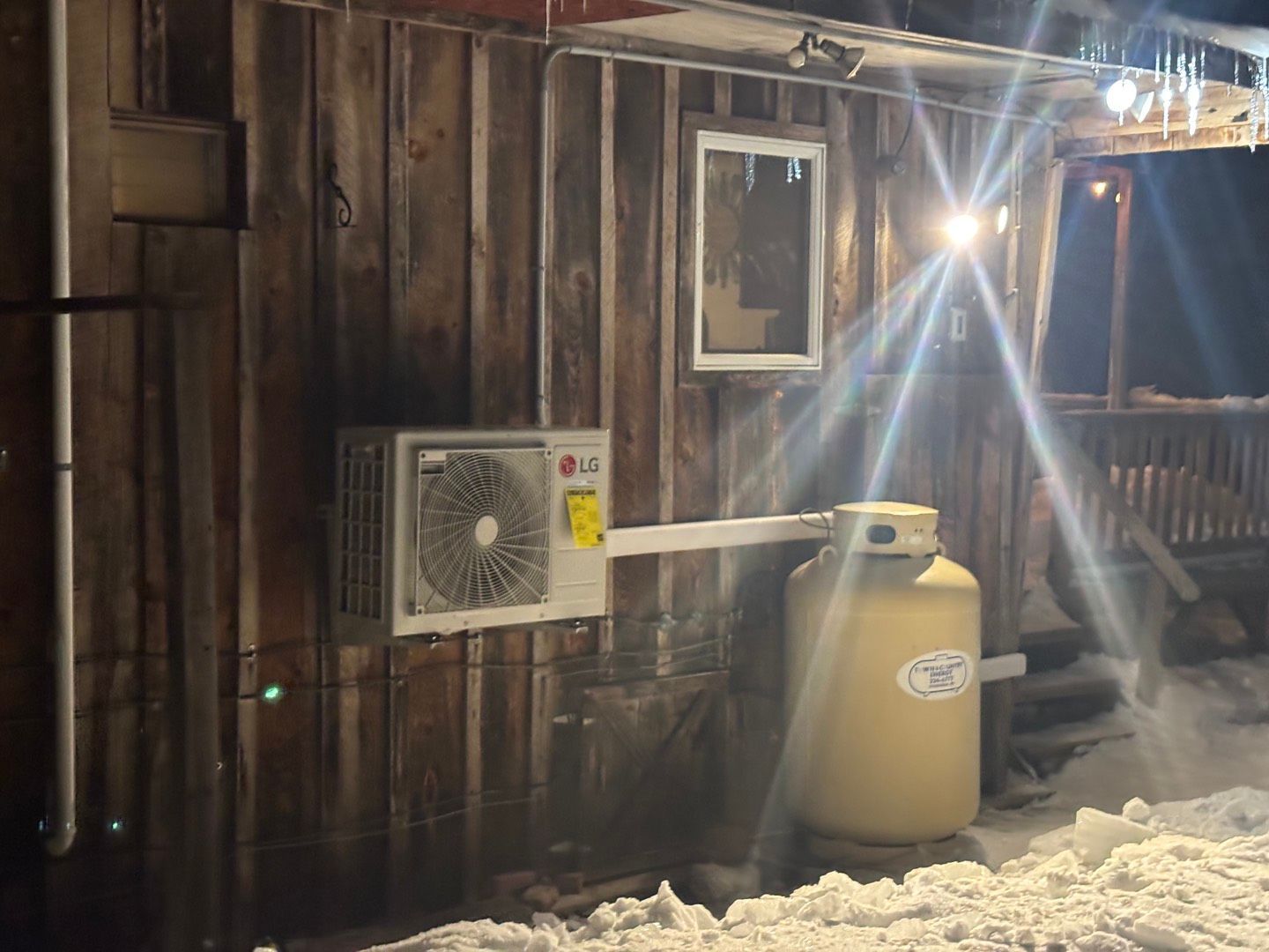 Snowy wooden house exterior with an air conditioner and a beige tank by the wall at night