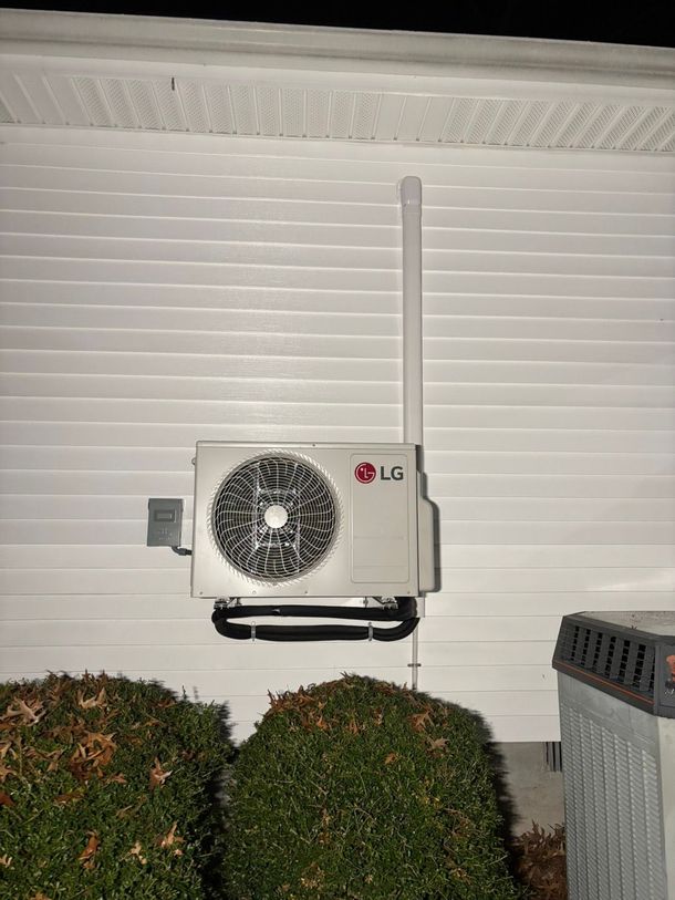 Wall-mounted LG air conditioner unit outside a house above trimmed bushes at night