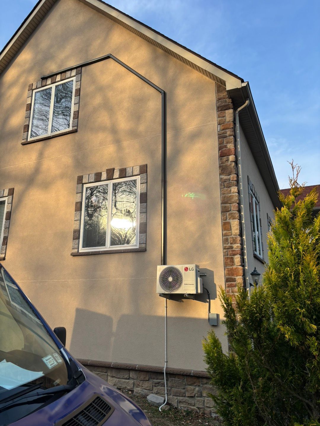 Sunlit beige house exterior with windows, a wall-mounted air conditioner, and a parked car edge in front