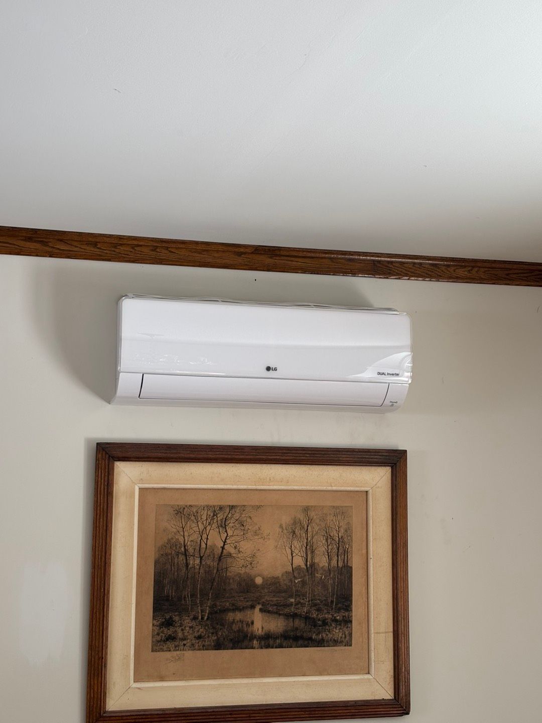 Wall-mounted white air conditioner above a framed landscape painting on a beige wall.