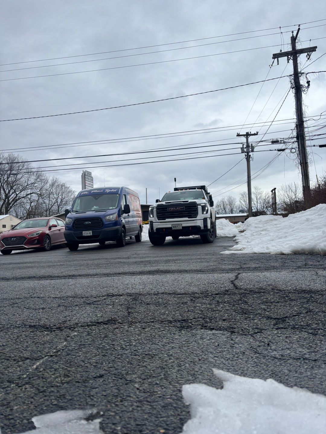 Cars lined up on a snowy roadside beneath power lines on an overcast day.