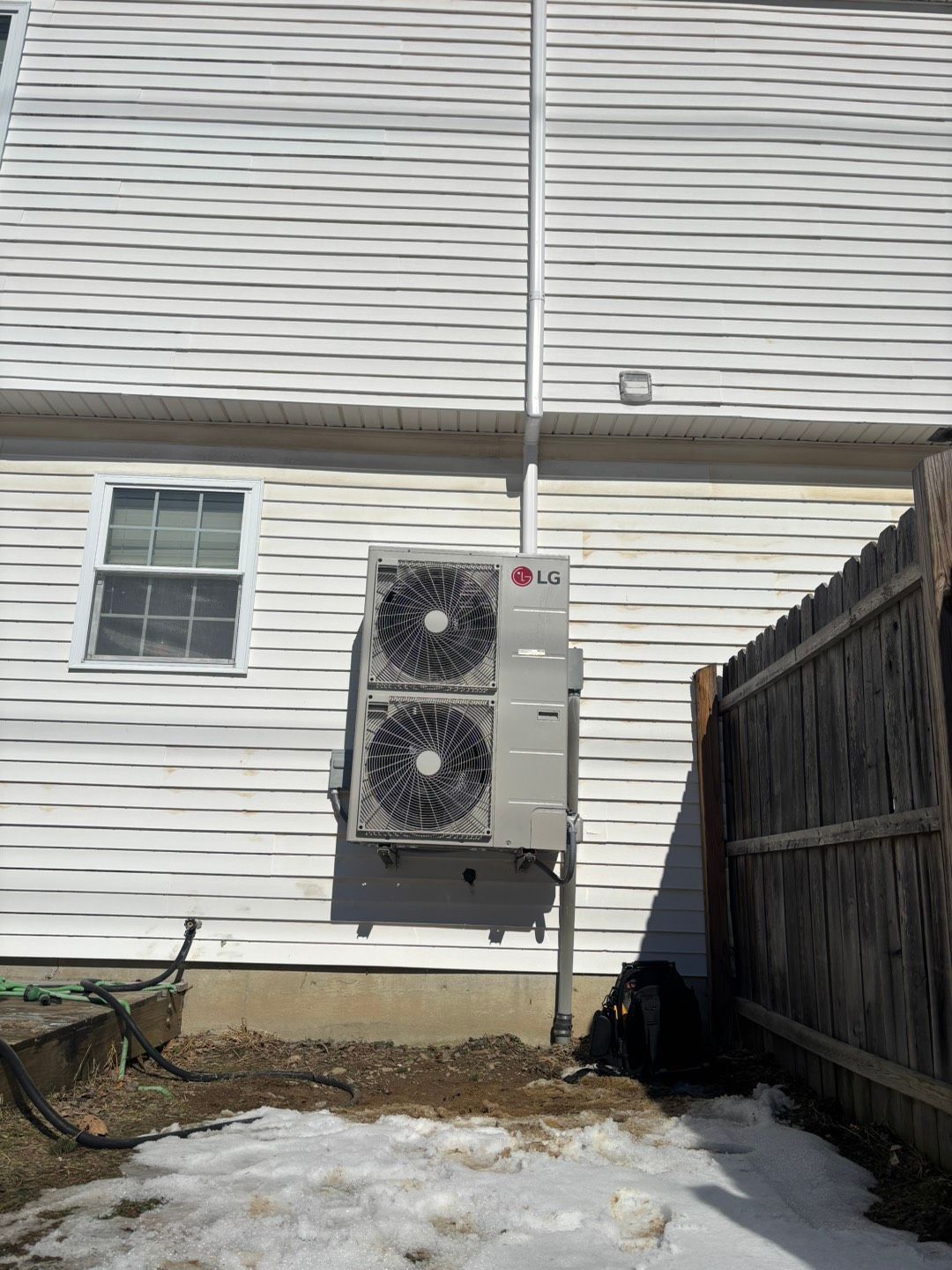 Two outdoor HVAC units mounted on a white house wall beside a fenced yard with patchy snow.