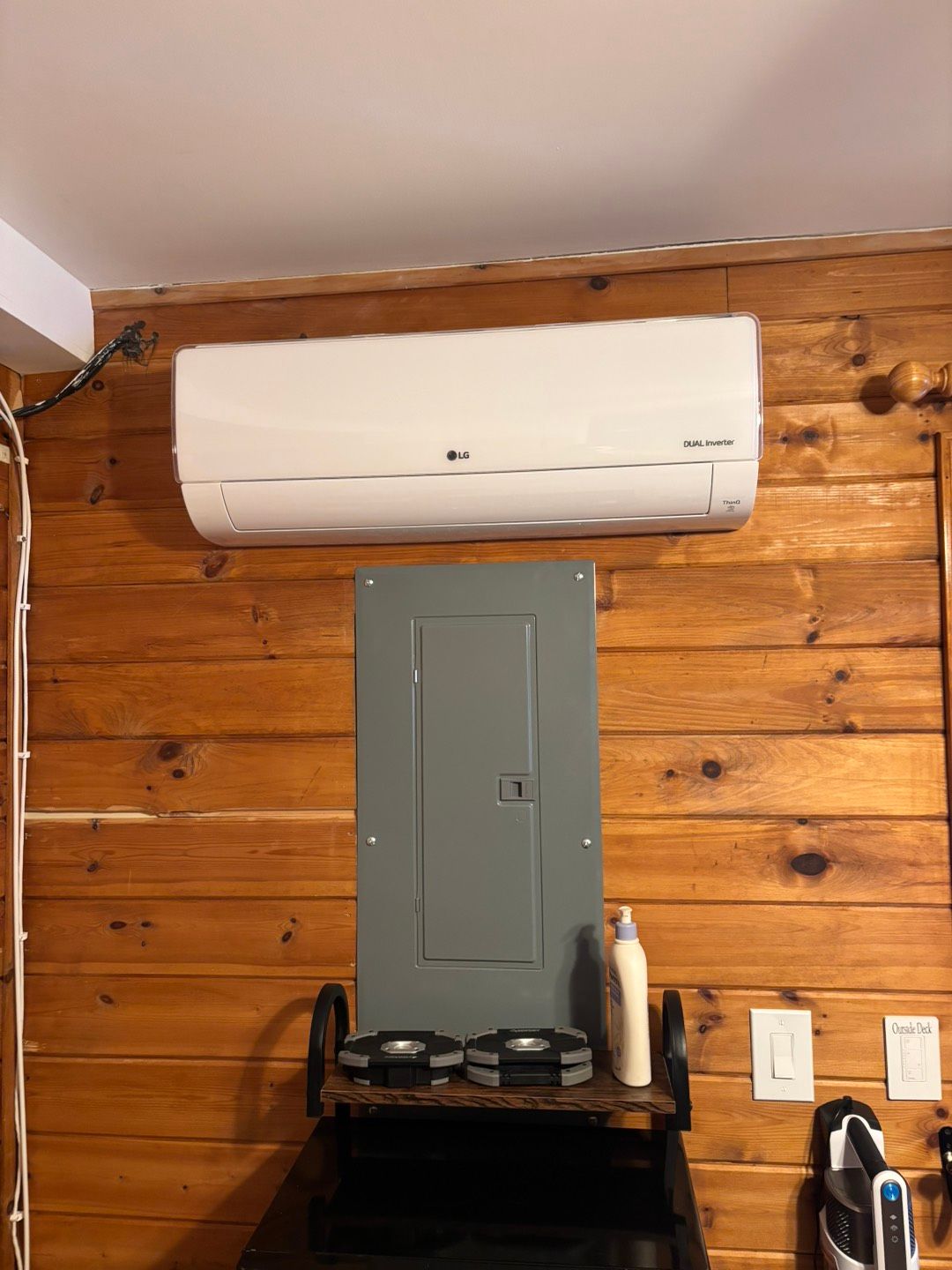 White wall-mounted air conditioner above a gray electrical panel on a wood-paneled wall.