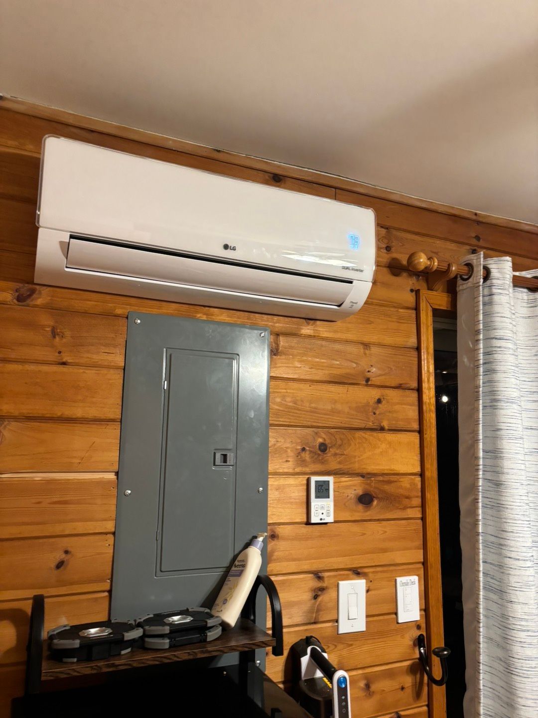 White wall-mounted AC above a gray electrical panel in a wood-paneled room with a ladder and wall controls