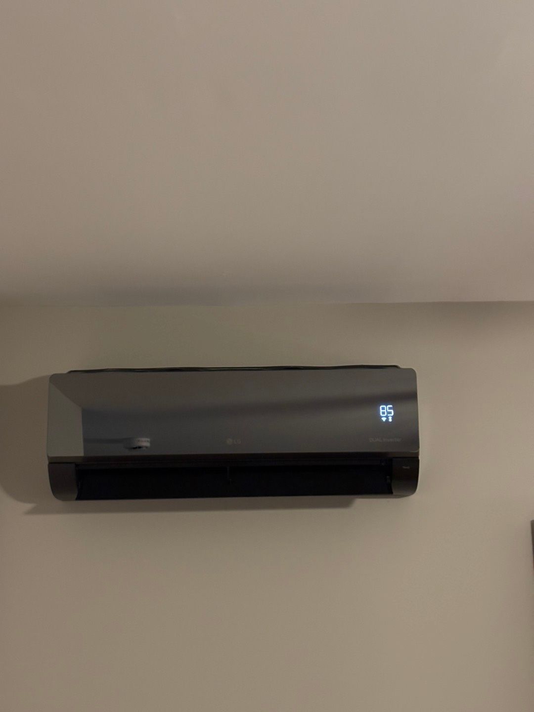 Wall-mounted gray air conditioner on a beige wall, lit display on the right side.