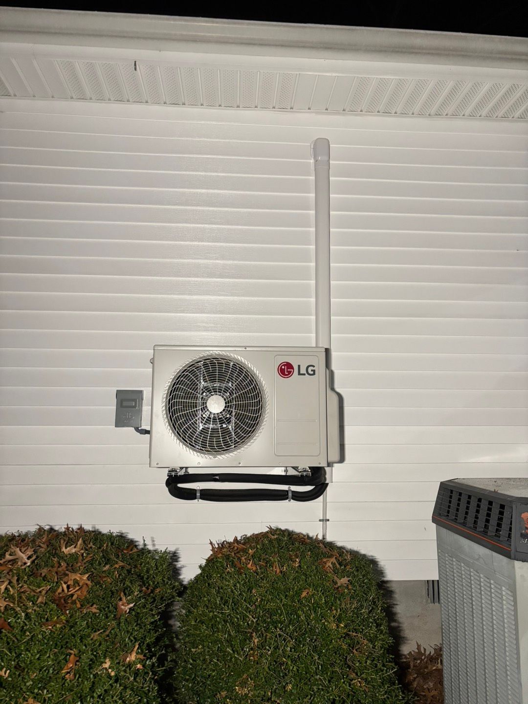 Outdoor wall-mounted LG air conditioner unit beside shrubs on a house exterior at night