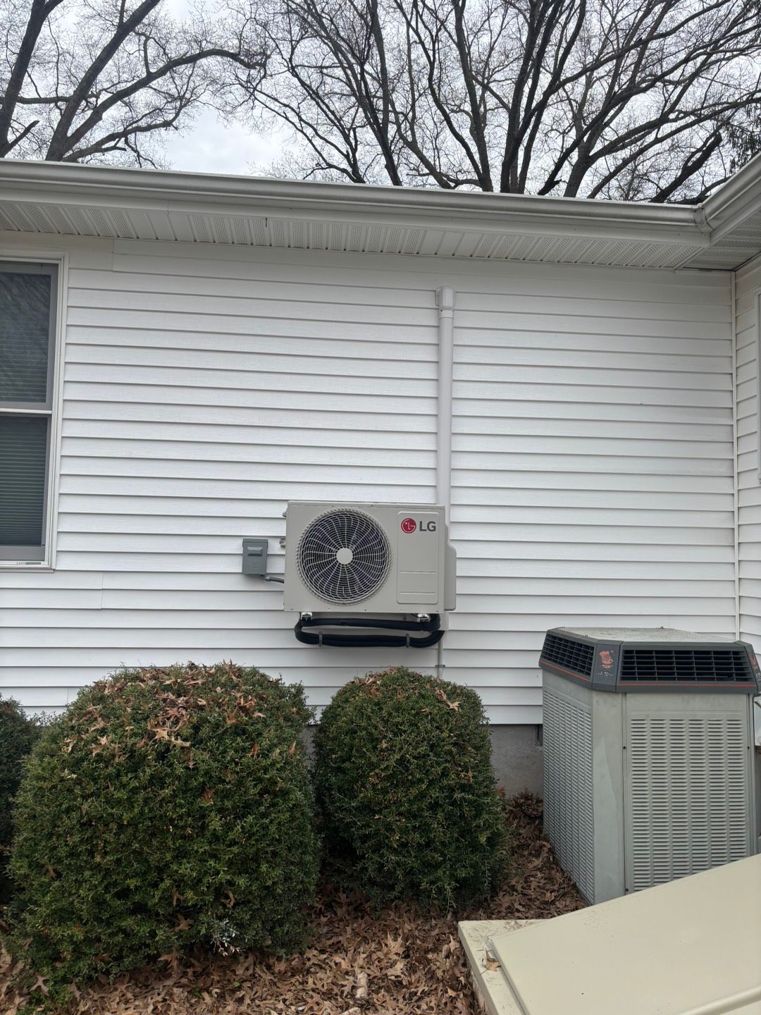 White house exterior with an outdoor HVAC unit mounted above two bushes and a larger AC unit on the right.
