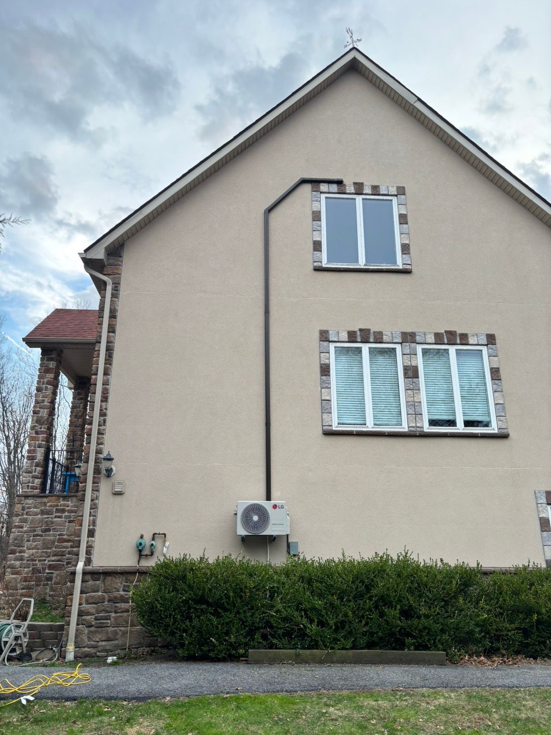 Side view of a beige house with white windows, an air conditioner, and shrubs along the base.